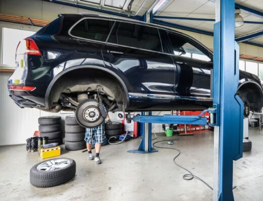 Auto in riparazione su un ponte sollevatore in un'officina meccanica, con un meccanico che lavora sul freno. Pneumatici impilati sullo sfondo.