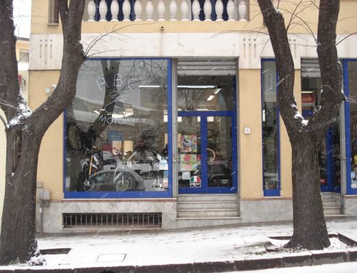 Foto della facciata di ABC Motors S.n.c., concessionario Aprilia, con motociclette visibili in vetrina durante una nevicata.