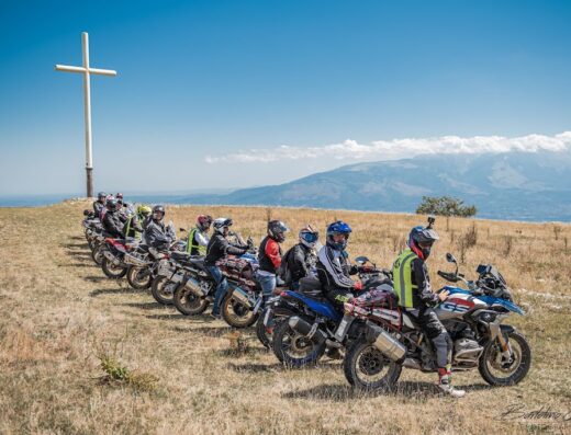 Gruppo di motociclisti Maxi Enduro in Abruzzo durante un tour avventura, con moto BMW GS, accessori da viaggio e sullo sfondo montagne e croce.