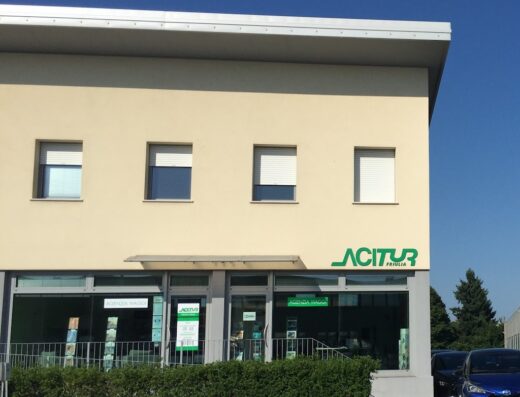 Foto dell'agenzia viaggi Acitur Friulia di Udintur Srl, con insegna verde ben visibile, vetrine con offerte viaggi e auto parcheggiate di fronte.