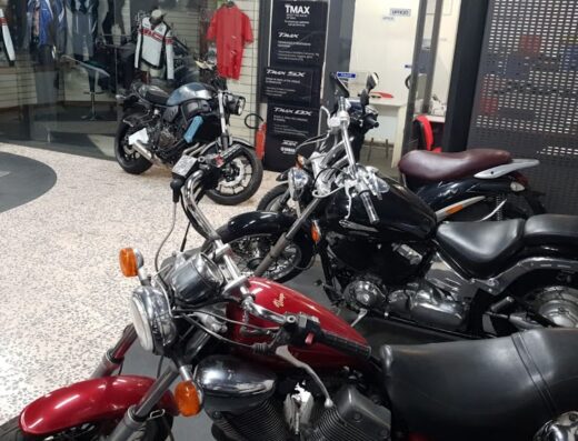 Diverse motorcycles showcased inside Adamoto dealership: A red cruiser in the foreground, a black cruiser behind, and a dark gray sport bike further back, surrounded by motorcycle apparel and TMAX promotional materials. These bikes are for sale or available for rent.