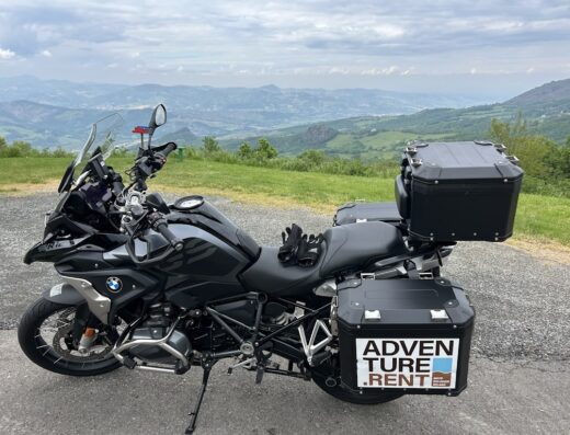 Noleggio moto adventure: BMW GS nera accessoriata con bauletti rigidi Adventure Rent, guanti sulla sella, sullo sfondo panorama collinare.