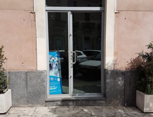 Agenzia Laika Viaggi: vetrina dell'agenzia viaggi con insegna Laika Viaggi, porta a vetro, cartellone pubblicitario e pavimentazione esterna.