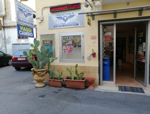 Ingresso dell'Agenzia Mufasa Viaggi e Turismo con insegne "Viaggi Cambio", "Mufasa Viaggi e Turismo Cambiavalute", piante di cactus ornamentali, auto parcheggiata e porta d'ingresso aperta.