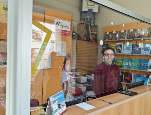 Un uomo dietro il bancone di un'agenzia viaggi Bramea, con Trenitalia sullo sfondo e guide turistiche esposte.
