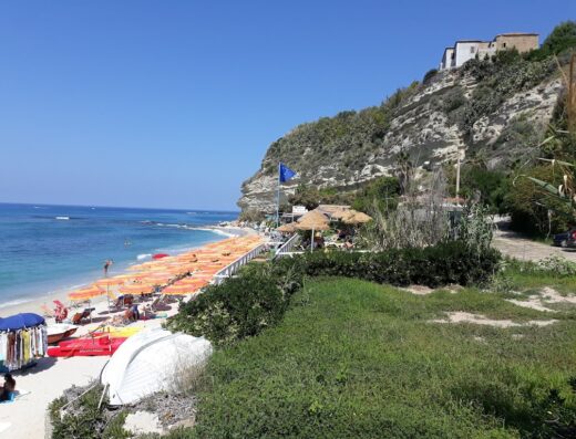 Spiaggia affollata con ombrelloni arancioni, mare cristallino e scogliera con vegetazione a Tropea, Calabria. Agenzia viaggi e turismo De Nicola Srl offre servizi per vacanze indimenticabili.