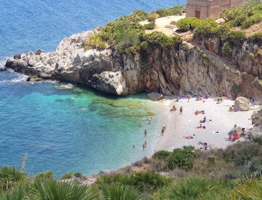 Vista panoramica di una spiaggia affollata in Sicilia con mare cristallino, scogliere rocciose e vegetazione mediterranea, ideale per viaggi e vacanze con Garit Viaggi.
