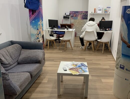 Interno dell'agenzia viaggi "I viaggi di Alice" con divano, tavolino con brochure, tappeto a tema aeroportuale e personale al lavoro.