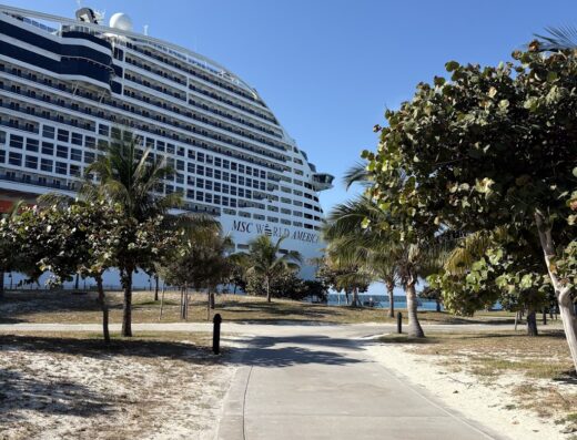 Una grande nave da crociera MSC World America sullo sfondo con una passerella di cemento in primo piano, alberi di palma e cielo azzurro. Perfetto per un'agenzia di viaggi che prepara le valigie.