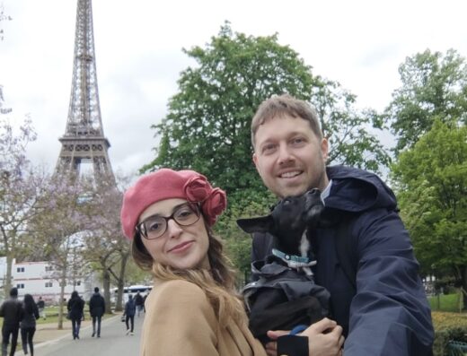 Una coppia sorridente con un cane vestito posa di fronte alla Torre Eiffel a Parigi, un viaggio indimenticabile con Kingston Travel.