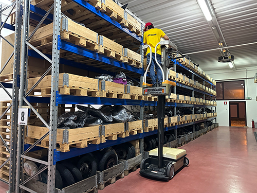 Immagine di un magazzino di AGMotoricambi con scaffali pieni di ricambi moto, un operatore su una piattaforma elevatrice preleva componenti. Vendita online di accessori e pezzi di ricambio per moto.