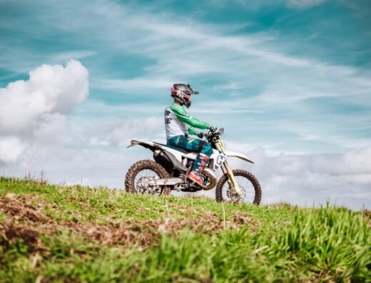 Pilota di Agrienduro su moto da cross Husqvarna, con abbigliamento tecnico e casco, su un terreno erboso con cielo azzurro nuvoloso sullo sfondo. Accessori e attrezzatura per motociclisti.