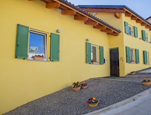 Agriostello delle Langhe: Facciata gialla con finestre verdi, vasi di fiori e vialetto di ghiaia. Un'accogliente struttura ricettiva nelle Langhe.