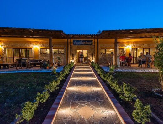 AgriQuartuccio b&b con piscina salata illuminata di sera, struttura in pietra con tavoli esterni e persone sedute, vialetto illuminato.