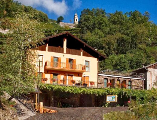 Agritur All'Olivo: Facciata esterna dell'agriturismo con balconi fioriti, situato in un paesaggio collinare con una chiesetta sullo sfondo.