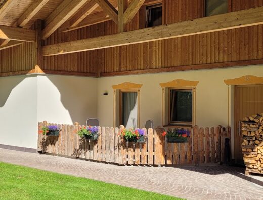 Agritur Majon Da Mont vista esterna con prato verde, recinzione in legno con fiori, tetto in legno e cielo azzurro.