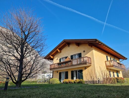 Agritur Val d'Adige vista esterna: edificio giallo a due piani con balconi in legno, finestre verdi, prato curato e albero spoglio.
