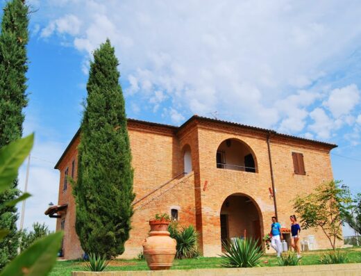 Agriturismo Farina: Facciata di una casa colonica in mattoni rossi con due cipressi, vaso in terracotta e due persone che camminano nel giardino.