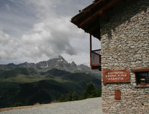 Agriturismo A Nostro Mizoun: facciata in pietra con balcone in legno, insegna e vista panoramica sulle montagne della Valle d'Aosta. Ideale per vacanze e turismo rurale.
