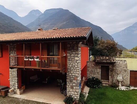 Agriturismo Al Picchio: Charming red farmhouse in Italian countryside with stone accents, balcony flowers, visible motorcycle parked under a covered area, and scenic mountain backdrop.