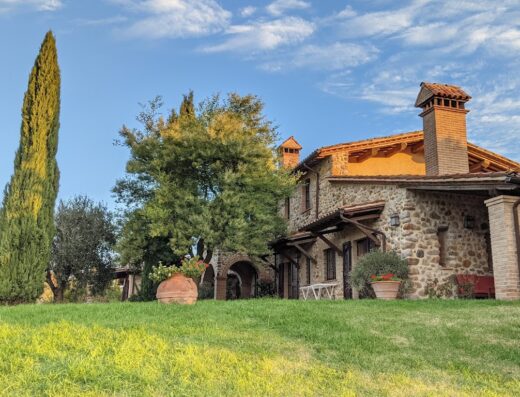 Agriturismo Belvedere Delle Crete: Tuscan stone farmhouse with terracotta roof and chimney, surrounded by green lawn and cypress trees under a blue sky.