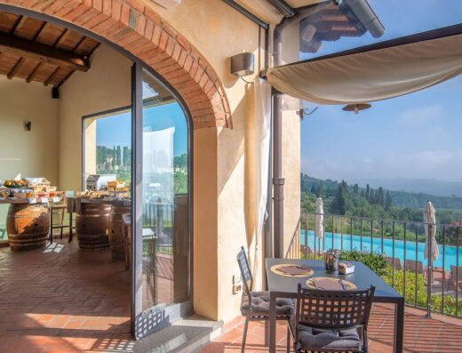 Terrazza panoramica dell'Agriturismo Borgo Divino con tavolo apparecchiato, piscina e vista sulla campagna toscana. Colazione a buffet con botti di legno.