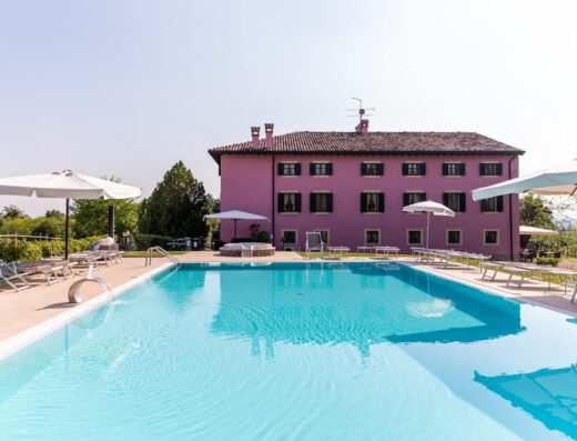 Agriturismo Cà Vecia vista piscina: Una grande piscina turchese domina il paesaggio, circondata da ombrelloni bianchi e lettini prendisole. Sullo sfondo, una casa colonica rosa con finestre chiuse offre un'atmosfera rilassante e accogliente.