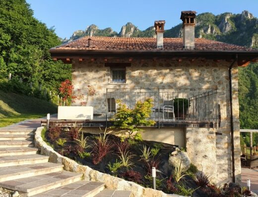Agriturismo Casarai vista esterna: casa in pietra con balcone, circondata da giardino curato e montagne sullo sfondo, tipico paesaggio italiano.