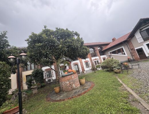 Cascina Argentera agriturismo vista esterna con pozzo decorativo, alberi e prato verde in una giornata nuvolosa.