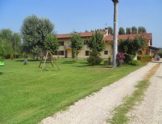 Agriturismo Cascina Gai: Veduta esterna della cascina con prato verde, alberi e vialetto di ghiaia in una giornata di sole.