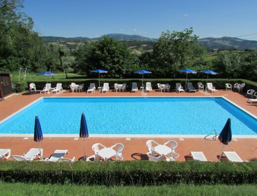 Piscina dell'Agriturismo Dea in Umbria, circondata da sedie a sdraio bianche, tavolini e ombrelloni blu, ideale per vacanze rilassanti.