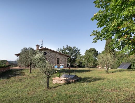 Agriturismo Dolce Sentire Assisi view of stone farmhouse with garden, olive trees, well, and solar panel under a clear blue sky.