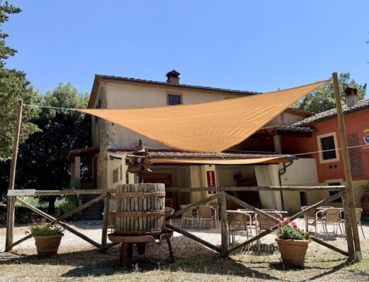 Agriturismo Fattoria La Prugnola in Toscana: vista esterna con tenda parasole, tino in legno decorativo e area relax con tavoli e sedie.