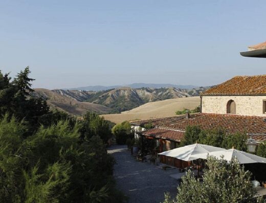 Panoramica dell'Agriturismo Fattoria Lischeto con vista sulle colline toscane, edifici in pietra con tetti rossi e ombrelloni bianchi.