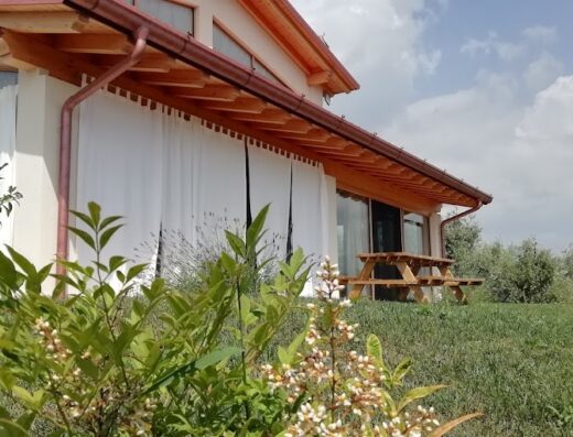 Agriturismo Fioralba: vista esterna della struttura con tendaggi bianchi, tavolo da picnic in legno e vegetazione rigogliosa in primo piano.