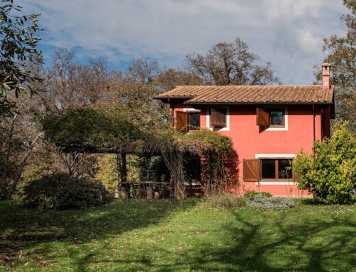Agriturismo Il Palombaio: Esterno di una casa colonica rossa con tetto in tegole, circondata da alberi e vegetazione lussureggiante.