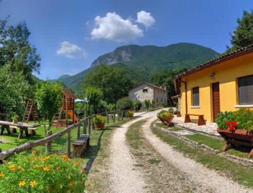 Agriturismo La Ferrera: A charming Italian farmhouse with a dirt road leading to stone buildings, a yellow house, a playground, and benches with flower boxes, set against a backdrop of lush green mountains under a blue sky with scattered clouds.