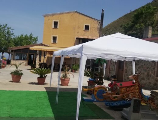 Agriturismo La Palombara: vista esterna con tenda bianca, edificio giallo, zona verde, giochi per bambini e auto parcheggiata.