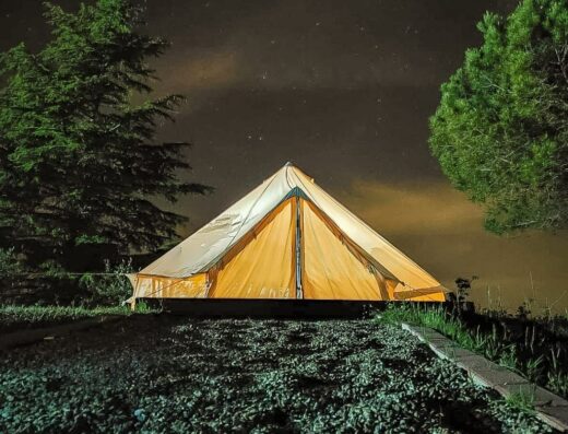 Tenda illuminata di notte all'Agriturismo La Taverna, con cielo stellato, alberi e terreno sassoso. Esperienza di campeggio e natura.