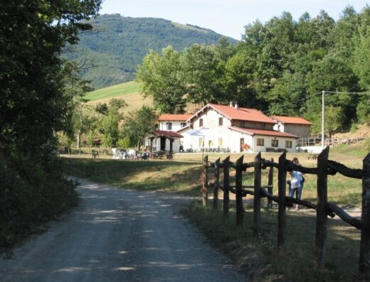 Agriturismo Le Carovane view featuring a gravel road leading to the building, with outdoor seating, surrounded by lush green trees and a mountain backdrop.