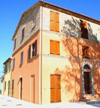 Agriturismo Le Piagge building exterior, featuring a renovated farmhouse with wooden shutters, stone accents, and warm color palette against a blue sky.