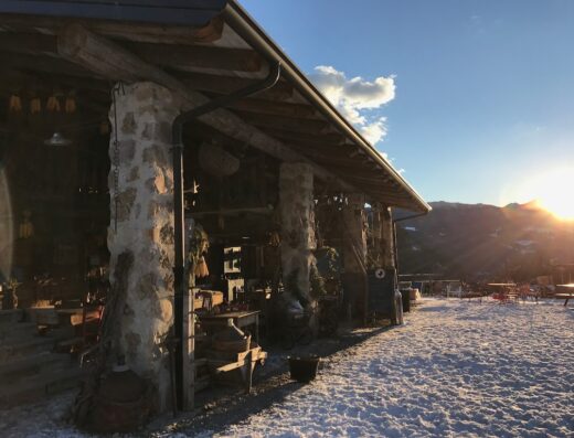 Agriturismo Malga Bassa: vista esterna della struttura in pietra con neve, tavoli all'aperto e vista sulle montagne al tramonto. Ideale per un viaggio e turismo esperienziale.