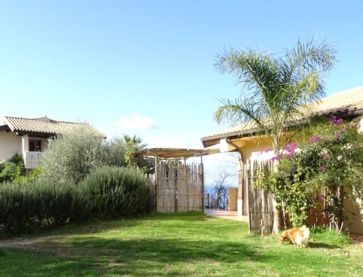 Agriturismo Pirapora: Vista di un giardino curato con prato verde, vegetazione mediterranea, edifici rurali e scorcio sul mare.