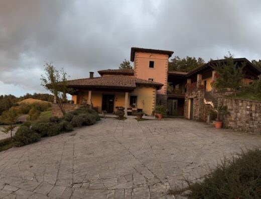 Agriturismo Podere Caselina vista esterna, edificio principale con torre, ampio piazzale in pietra, colline sullo sfondo al tramonto.