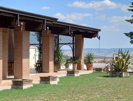 Agriturismo Tempa Bianca: View of the building's architecture with brick columns, plants in pots, a green lawn, and a distant landscape under a blue, cloudy sky.