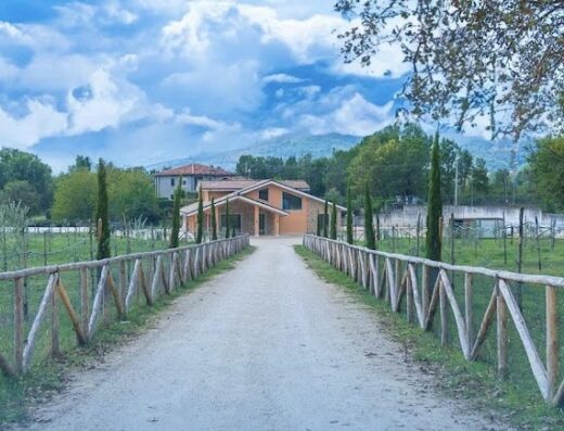 Strada sterrata che conduce all'Agriturismo Tenuta Santa Barbara, con recinzione in legno e alberi di cipresso, in una giornata di sole con cielo azzurro e nuvole.