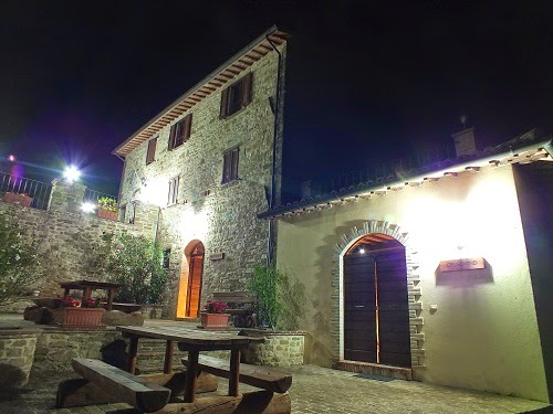 Agriturismo Val di Boccio at night. Stone building with wooden picnic tables and benches outside. Warm light glows from arched doorways.
