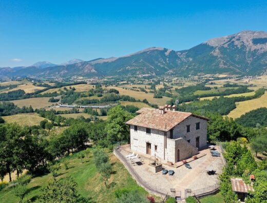 Agriturismo Villa Dama: Aerial view of stone farmhouse with tiled roof, surrounded by rolling hills and mountains in Italy.