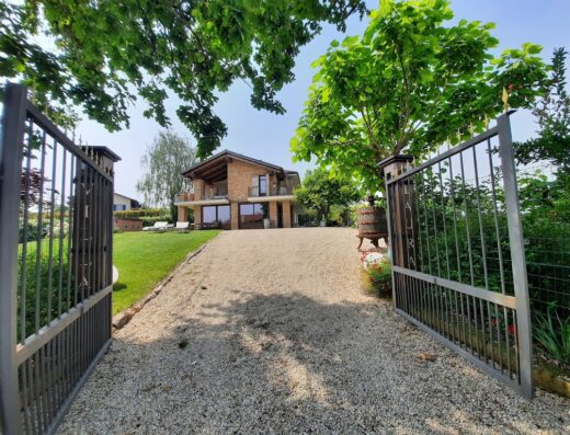 Agriturismo Villa Laura: cancello d'ingresso aperto che rivela la splendida villa con vialetto di ghiaia, prato verde e alberi rigogliosi.