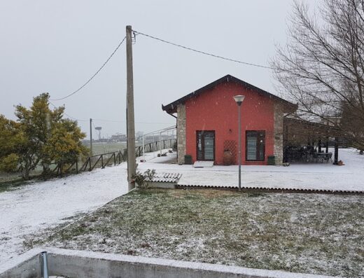 Agriturismo Zarita in inverno: Casale rosso immerso nella neve, con prato innevato, albero spoglio e cielo grigio. Ideale per un soggiorno rilassante e autentico.
